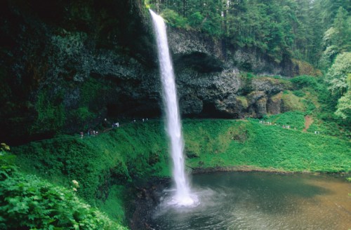 A tall waterfall cascades into a serene pool, surrounded by lush greenery and rocky cliffs.