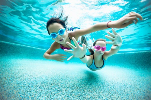 Two children swim underwater, wearing colorful goggles, reaching out with their hands in a bright blue pool.
