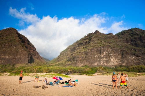 A sandy beach scene with people relaxing and playing, surrounded by lush green mountains and a blue sky.
