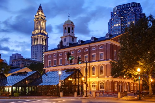 Historic building illuminated at dusk, with a clock tower and modern skyscrapers in the background.