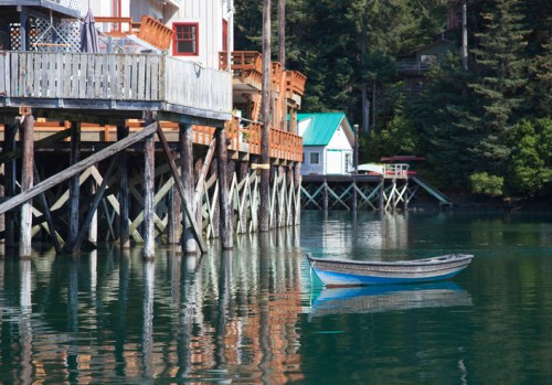 A small blue boat floats in calm water near a wooden pier surrounded by trees and colorful buildings.