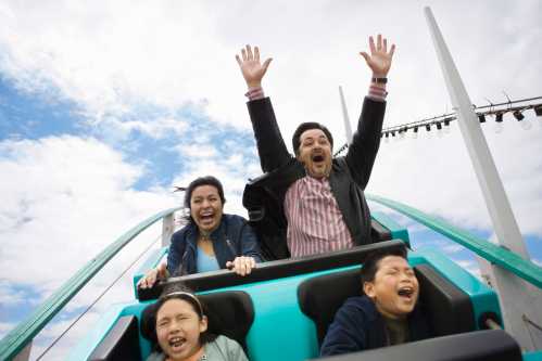 A family enjoys a roller coaster ride, laughing and raising their hands in excitement against a cloudy sky.