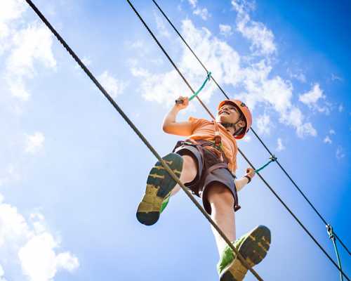 A child in an orange helmet climbs a rope high against a blue sky with fluffy clouds.