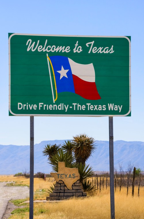 Welcome to Texas sign featuring the Texas flag, with a stone Texas shape and desert landscape in the background.