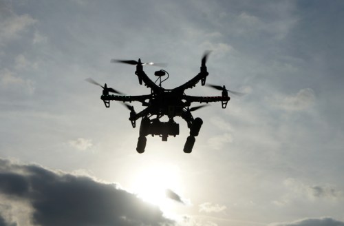 A silhouette of a drone flying against a cloudy sky with the sun partially visible in the background.