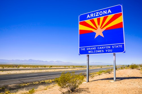 A welcome sign for Arizona featuring the state flag and the text "The Grand Canyon State Welcomes You" against a clear sky.