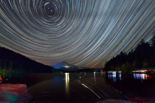Star trails swirl above a serene lake, reflecting trees and a mountain in the background, creating a tranquil night scene.