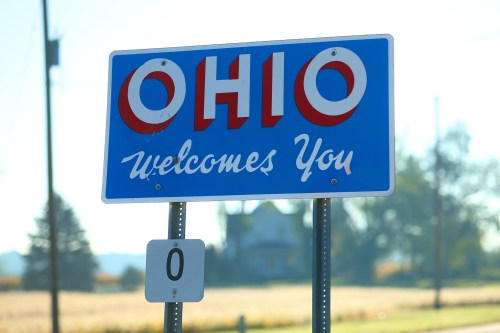 A blue sign that reads "Ohio Welcomes You" with a white background and red lettering, set against a rural landscape.