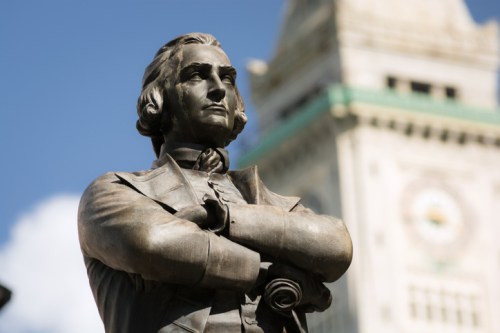 A close-up of a bronze statue of a historical figure with arms crossed, set against a blurred clock tower background.