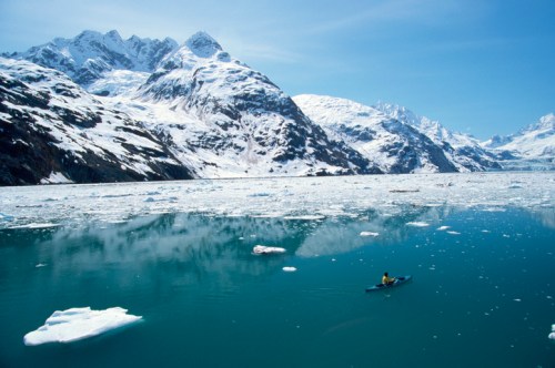 A kayaker paddles through icy waters surrounded by snow-capped mountains and floating ice in a serene landscape.