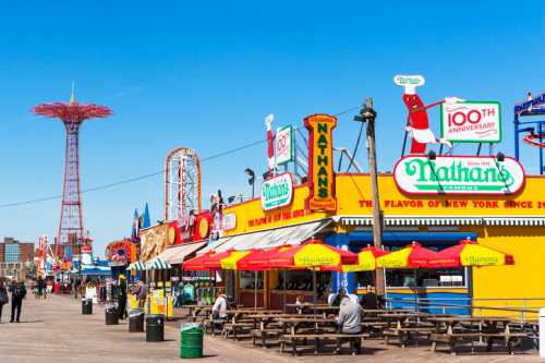 Colorful boardwalk scene featuring Nathan's Famous, amusement rides, and a clear blue sky.