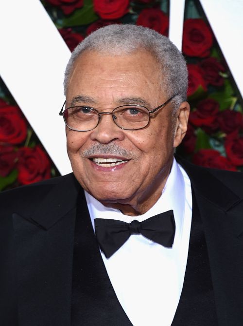 A smiling older man with glasses and gray hair, wearing a tuxedo, stands in front of a backdrop of red roses.