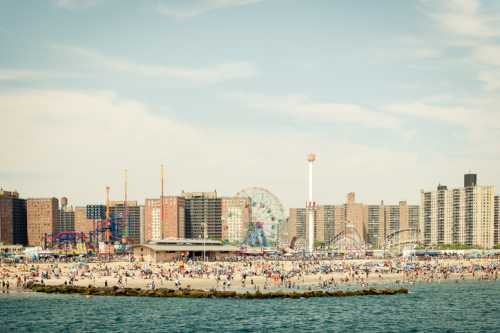 A bustling beach scene with a boardwalk, amusement rides, and crowds against a backdrop of city buildings.