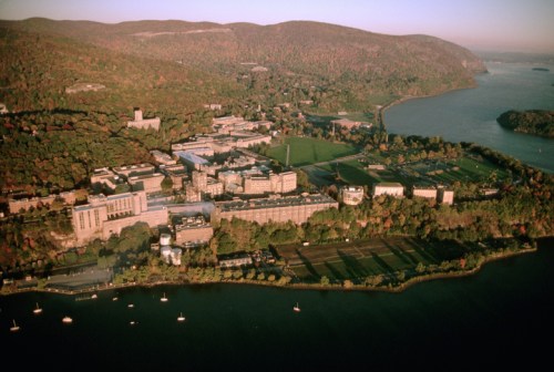 Aerial view of a riverfront campus surrounded by autumn foliage and mountains, with boats on the water.