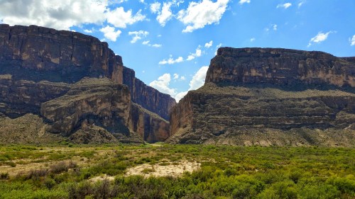 A scenic view of rugged cliffs and canyons under a bright blue sky with scattered clouds, surrounded by green vegetation.