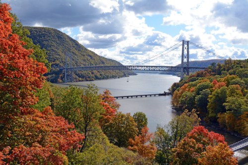 A scenic view of a river with a bridge, surrounded by vibrant autumn foliage and rolling hills under a cloudy sky.