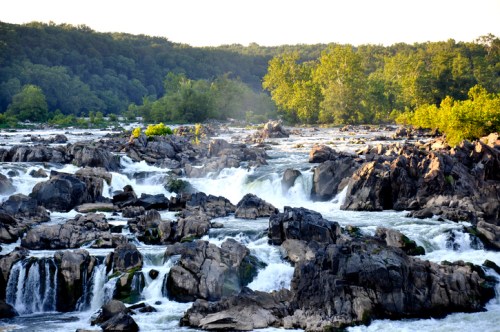 A scenic view of a waterfall cascading over rocky terrain, surrounded by lush green trees and a clear blue sky.