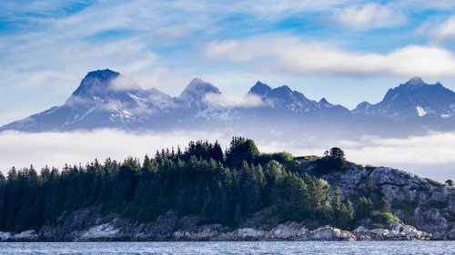 A serene landscape featuring a forested island in the foreground and majestic, snow-capped mountains in the background.
