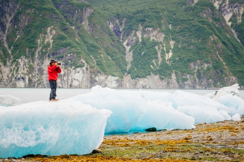 A person in a red jacket stands on an ice floe, photographing the scenic mountains and water in the background.