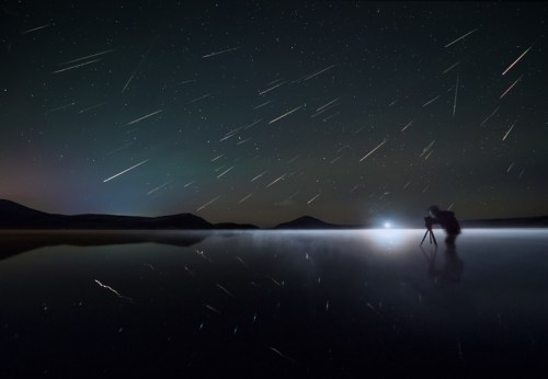 A photographer captures a starry night with shooting stars reflected in a calm lake, surrounded by mountains.