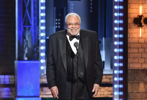 A smiling man in a tuxedo stands at a microphone on stage, with a lit backdrop and audience in the background.