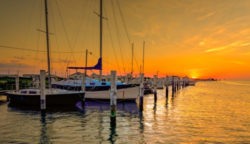 Sunset over a calm marina with sailboats docked along wooden piers, reflecting warm colors on the water.
