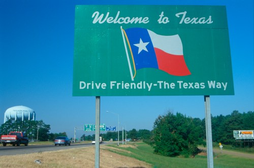 A green welcome sign for Texas featuring the state flag, with cars and a water tower in the background.