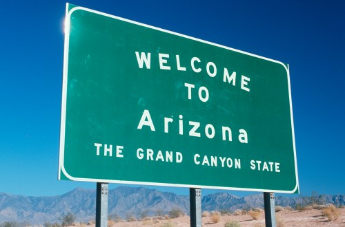 Green welcome sign for Arizona, stating "Welcome to Arizona, The Grand Canyon State," with mountains in the background.