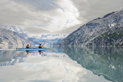 Two kayakers paddle on a calm lake, surrounded by majestic mountains and a cloudy sky, reflecting on the water's surface.