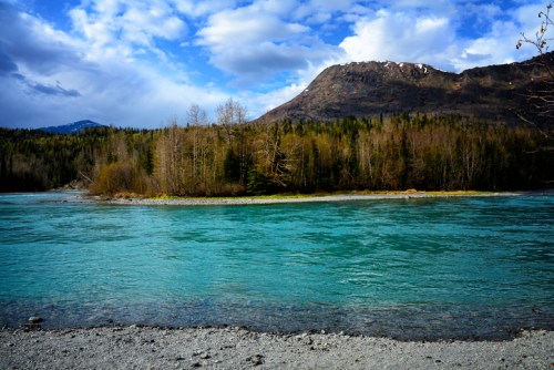 A serene river flows through a landscape of trees and mountains under a partly cloudy sky.