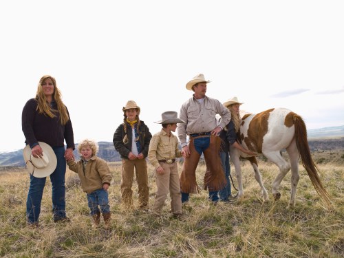 A family in cowboy attire stands in a grassy field with a horse, set against a cloudy sky and distant mountains.