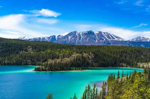 A stunning landscape featuring a turquoise lake surrounded by lush forests and snow-capped mountains under a blue sky.