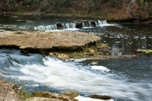 A serene river scene with gentle waterfalls cascading over rocky ledges surrounded by lush greenery.