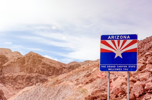 A welcome sign for Arizona featuring a star and rays, set against a rocky landscape and blue sky.