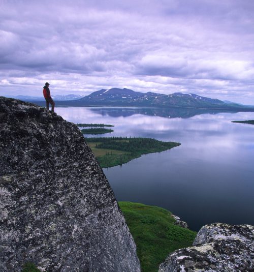 A person stands on a rocky ledge overlooking a serene lake and distant mountains under a cloudy sky.