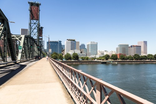 A view of a bridge with a walkway, overlooking a river and a city skyline on a clear day.