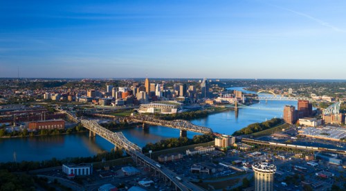 Aerial view of a city skyline with bridges over a river, surrounded by greenery and urban buildings under a clear blue sky.