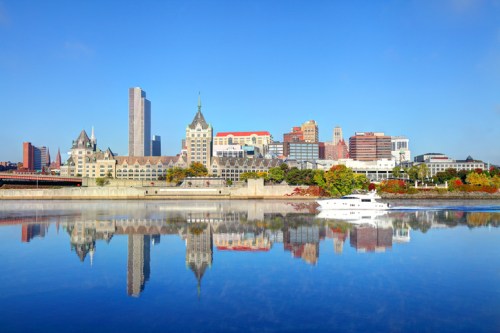 A clear blue sky reflects on a river, showcasing a city skyline with modern and historic buildings.