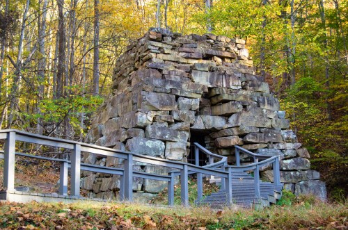 Stone structure surrounded by trees, with a wooden railing and steps leading to its entrance. Autumn foliage visible.