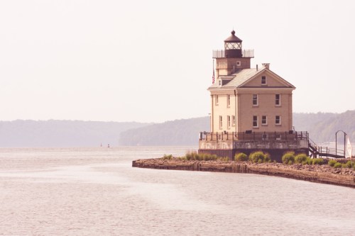 A vintage lighthouse stands on a riverbank, surrounded by calm water and distant hills under a soft, hazy sky.