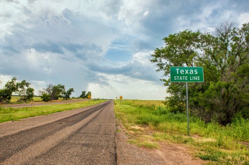 A road sign indicating the Texas state line, with a rural landscape and cloudy sky in the background.