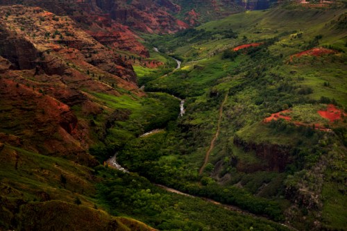 A lush green valley with a winding river, surrounded by red and brown rocky cliffs under a cloudy sky.