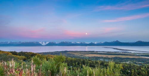 A serene landscape featuring mountains, a calm lake, and a colorful sky at dusk, with lush greenery in the foreground.
