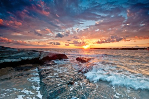 A vibrant sunset over the ocean, with colorful clouds and waves crashing against rocky shorelines.