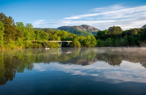 A serene lake surrounded by lush greenery and mountains, with mist rising from the water under a clear blue sky.