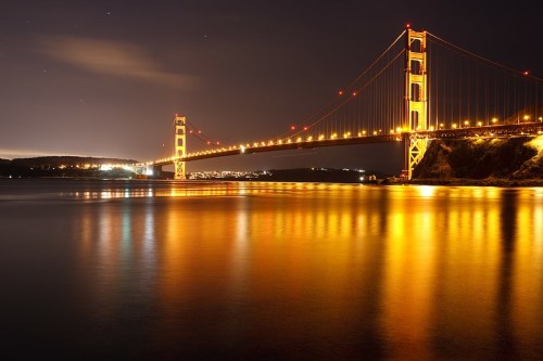 Golden Gate Bridge illuminated at night, reflecting in calm waters with a starry sky above.