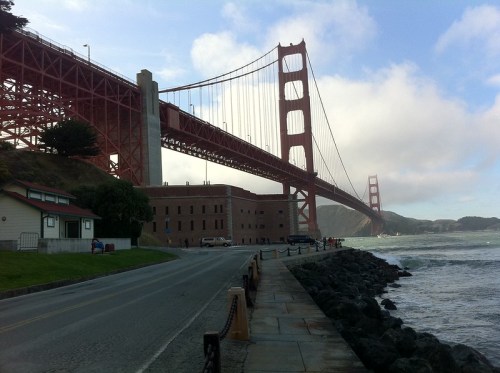 Golden Gate Bridge spans across a bay, with a rocky shoreline and a road lined with trees and buildings in the foreground.