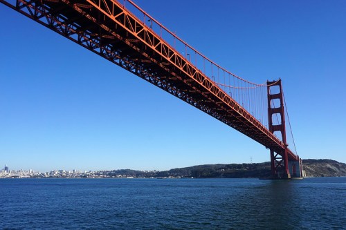 Golden Gate Bridge spans over a calm bay, with a clear blue sky and distant city skyline in the background.