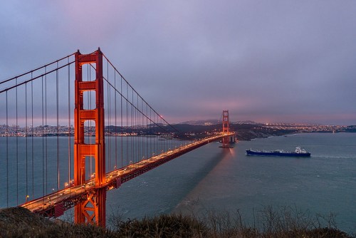 Golden Gate Bridge at dusk, with a ship passing beneath and city lights twinkling in the background.