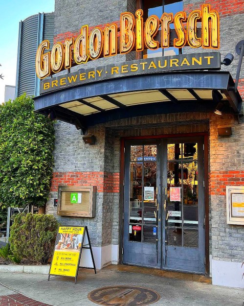 Entrance of Gordon Biersch Brewery-Restaurant with a sign and greenery around the door.